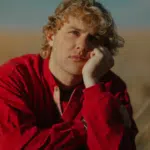 A pensive individual with curly hair resting their chin on their hand in a field, listening to fresh new music.