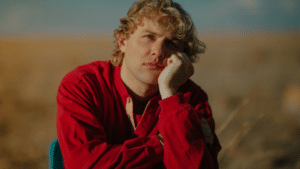 A pensive individual with curly hair resting their chin on their hand in a field, listening to fresh new music.