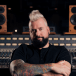 A man with a mohawk and tattoos sitting confidently in a music studio with mixing boards and speakers in the background, working on fresh new music.