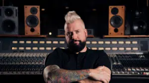 A man with a mohawk and tattoos sitting confidently in a music studio with mixing boards and speakers in the background, working on fresh new music.