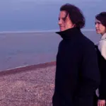 Two people in dark clothing walk along a pebble beach by the sea at dusk, looking away from the camera, discussing fresh new music.