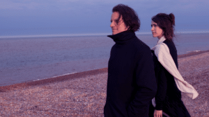 Two people in dark clothing walk along a pebble beach by the sea at dusk, looking away from the camera, discussing fresh new music.