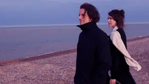 Two people in dark clothing walk along a pebble beach by the sea at dusk, looking away from the camera, discussing fresh new music.