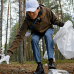 A person in a cap and gloves picks up eco-friendly litter in a forest with another individual visible in the background.