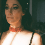 A woman in a beaded dress and choker poses indoors, half her face illuminated by natural light.
