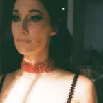 A woman in a beaded dress and choker poses indoors, half her face illuminated by natural light.