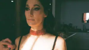 A woman in a beaded dress and choker poses indoors, half her face illuminated by natural light.