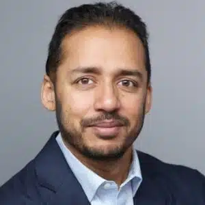 Portrait of a smiling man with light brown complexion, wearing a dark suit and light blue shirt, against a gray background.