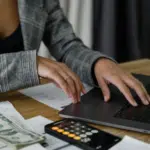 A person working on a laptop with documents, a calculator, and cash on the desk, indicating financial management or budgeting tasks.