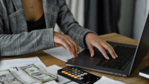 A person working on a laptop with documents, a calculator, and cash on the desk, indicating financial management or budgeting tasks.