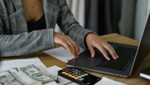 A person working on a laptop with documents, a calculator, and cash on the desk, indicating financial management or budgeting tasks.