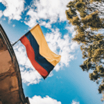 Colombian flag waving against a bright blue sky, framed by tree branches and an architectural element of a BIME building.
