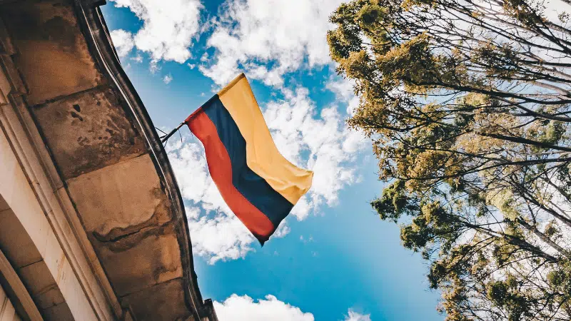 Colombian flag waving against a bright blue sky, framed by tree branches and an architectural element of a BIME building.