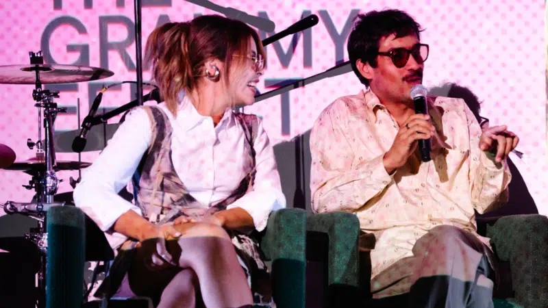 Two people are seated on stage, smiling and talking. One holds a microphone, engaging the audience in an UNSIN conversation. A drum set is visible against a pink backdrop, adding a vibrant touch to the lively scene.