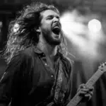 A long-haired guitarist passionately plays and sings on stage under bright lights, channeling emotion and supporting mental health through a powerful live music performance.