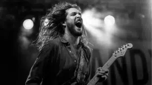A long-haired guitarist passionately plays and sings on stage under bright lights, channeling emotion and supporting mental health through a powerful live music performance.