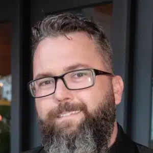 Portrait of a smiling man with a beard and glasses, wearing a black shirt, against a dark blurred background.