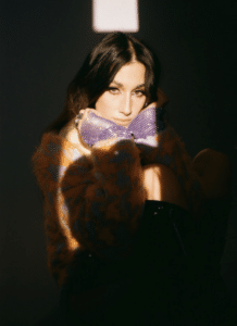 A woman poses in shadow, wearing a colorful fur coat and gloves, looking intently at the camera.
