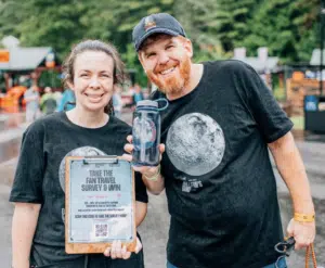 Two smiling adults, one holding an eco-friendly water bottle and the other holding a clipboard with a survey form, standing outdoors at an event.