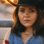 A person wearing a cowboy hat and denim clothing with a serious expression at twilight.