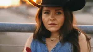 A person wearing a cowboy hat and denim clothing with a serious expression at twilight.