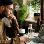 A focused man in a beanie and glasses works on a laptop at a wooden table in a cozy, plant-filled room.