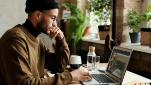 A focused man in a beanie and glasses works on a laptop at a wooden table in a cozy, plant-filled room.