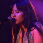 A woman of AAPI descent singing into a microphone on a stage, illuminated by purple lighting, with eyes closed and expressive features.