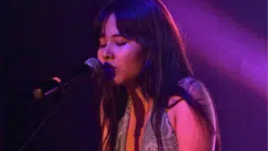 A woman of AAPI descent singing into a microphone on a stage, illuminated by purple lighting, with eyes closed and expressive features.