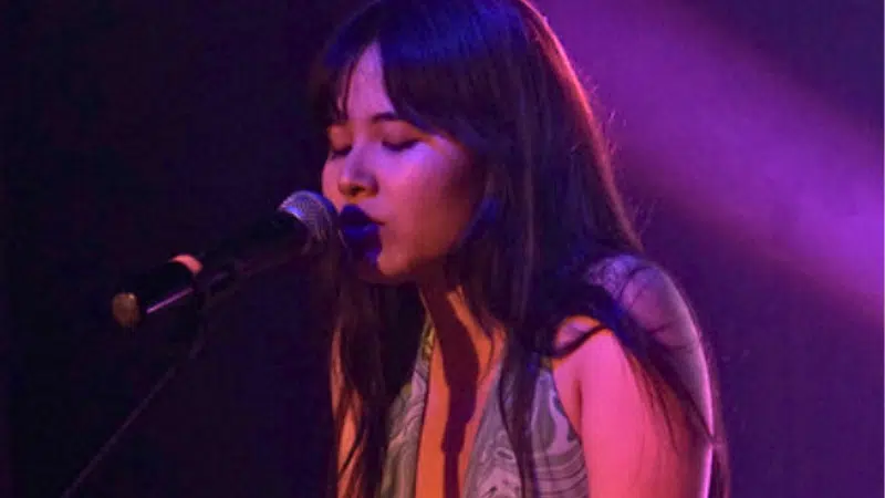 A woman of AAPI descent singing into a microphone on a stage, illuminated by purple lighting, with eyes closed and expressive features.