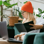 Woman in orange beanie using a laptop and reading a notebook on a green sofa, surrounded by indoor plants.