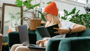 Woman in orange beanie using a laptop and reading a notebook on a green sofa, surrounded by indoor plants.