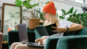 Woman in orange beanie using a laptop and reading a notebook on a green sofa, surrounded by indoor plants.