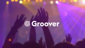 Hands raised at a music promotion concert with colorful stage lights, featuring the logo "groover" foregrounded.