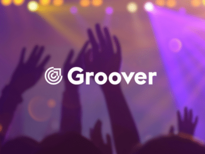 Hands raised at a music promotion concert with colorful stage lights, featuring the logo "groover" foregrounded.