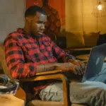 A person in a red plaid shirt and jeans sits in a chair, using a laptop. Headphones rest on a nearby table in the dimly lit room with modern decor, perfect for catching up on SplitShare projects.