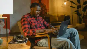 A person in a red plaid shirt and jeans sits in a chair, using a laptop. Headphones rest on a nearby table in the dimly lit room with modern decor, perfect for catching up on SplitShare projects.