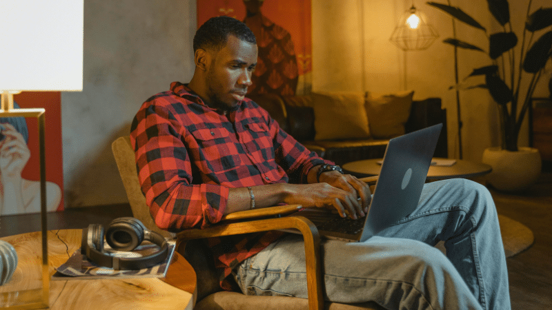 A person in a red plaid shirt and jeans sits in a chair, using a laptop. Headphones rest on a nearby table in the dimly lit room with modern decor, perfect for catching up on SplitShare projects.