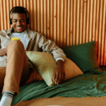 A person wearing headphones, sitting on a bed with wooden paneling in the background, smiles while looking at a yellow smartphone. They appear to be enjoying some music streams. The lamp on the bedside table is lit.