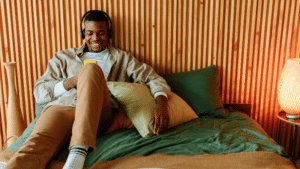 A person wearing headphones, sitting on a bed with wooden paneling in the background, smiles while looking at a yellow smartphone. They appear to be enjoying some music streams. The lamp on the bedside table is lit.