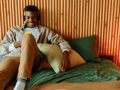 A person wearing headphones, sitting on a bed with wooden paneling in the background, smiles while looking at a yellow smartphone. They appear to be enjoying some music streams. The lamp on the bedside table is lit.