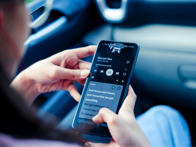 Person holding a smartphone inside a car, streaming music with visible lyrics on the screen, captivated by an unseen audience through their device.