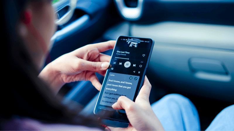 Person holding a smartphone inside a car, streaming music with visible lyrics on the screen, captivated by an unseen audience through their device.