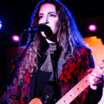 A woman with long curly hair plays an electric guitar and sings into a microphone on stage with colored lights in the background, conquering her imposter syndrome with every powerful note.