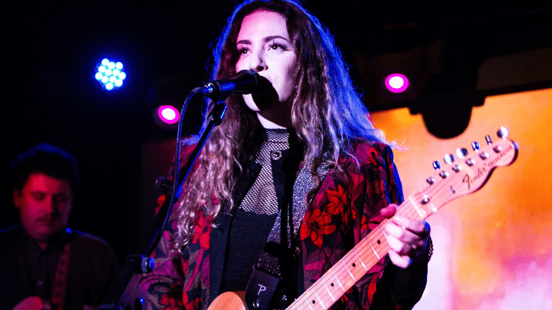 A woman with long curly hair plays an electric guitar and sings into a microphone on stage with colored lights in the background, conquering her imposter syndrome with every powerful note.