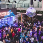 A crowded indoor event with a "INDIEWEEK" banner, showcasing various people socializing under a disco ball amid purple lighting. The event features a bar area to one side.