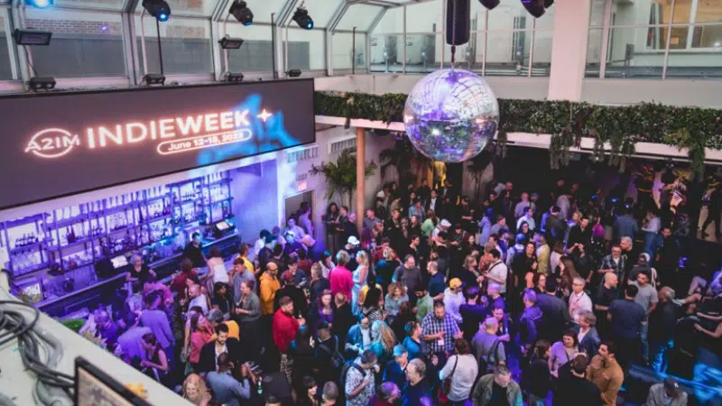 A crowded indoor event with a "INDIEWEEK" banner, showcasing various people socializing under a disco ball amid purple lighting. The event features a bar area to one side.