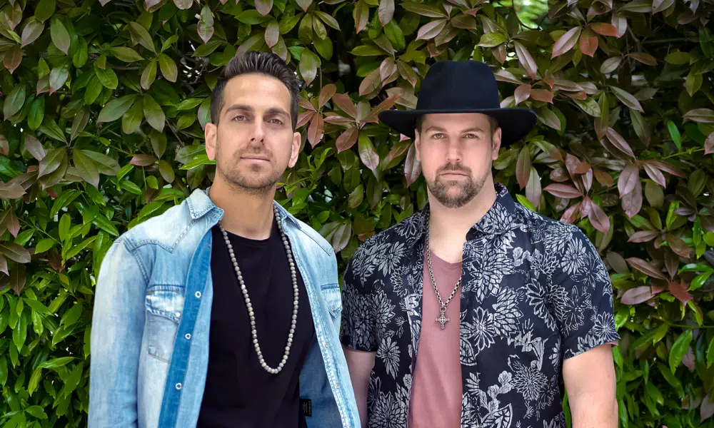 Two men standing in front of a leafy green background, ready to share their new music. One wears a denim shirt and beaded necklace, while the other sports a black hat and patterned shirt.