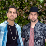 Two men standing in front of a leafy green background, ready to share their new music. One wears a denim shirt and beaded necklace, while the other sports a black hat and patterned shirt.