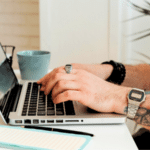 A person with tattooed arms streaming content on a laptop at a desk with a notebook, coffee mug, and plant nearby.